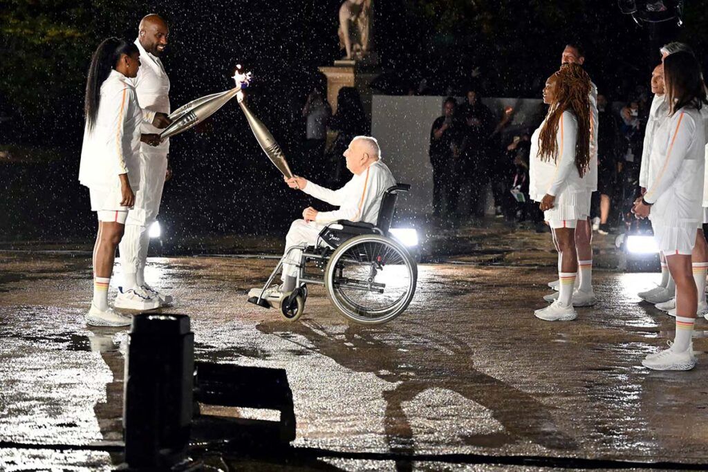 Nosilac baklje, bivši francuski biciklista Charles Coste , predaje olimpijsku vatru bivšoj francuskoj sprinterki Marie-José Perec i francuskom džudisti Teddyju Rineru tokom ceremonije otvaranja Olimpijskih igara